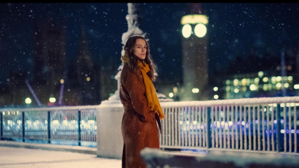 Keira Knightley stands on a snowy bridge at night in a scene from Netflix’s spy thriller Black Doves. Dressed in a warm coat and scarf, she gazes pensively into the distance, with city lights and falling snow creating a moody, atmospheric backdrop.